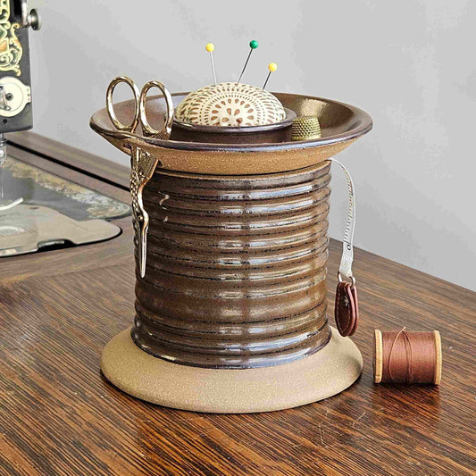 A ceramic sewing organizer caddy shaped like an oversized spool with removable lid, scissor holder and pincushion topper. It is placed on a wooden surface next to an old sewing machine with a few sewing accessories like scissors and bobbins stored on it.