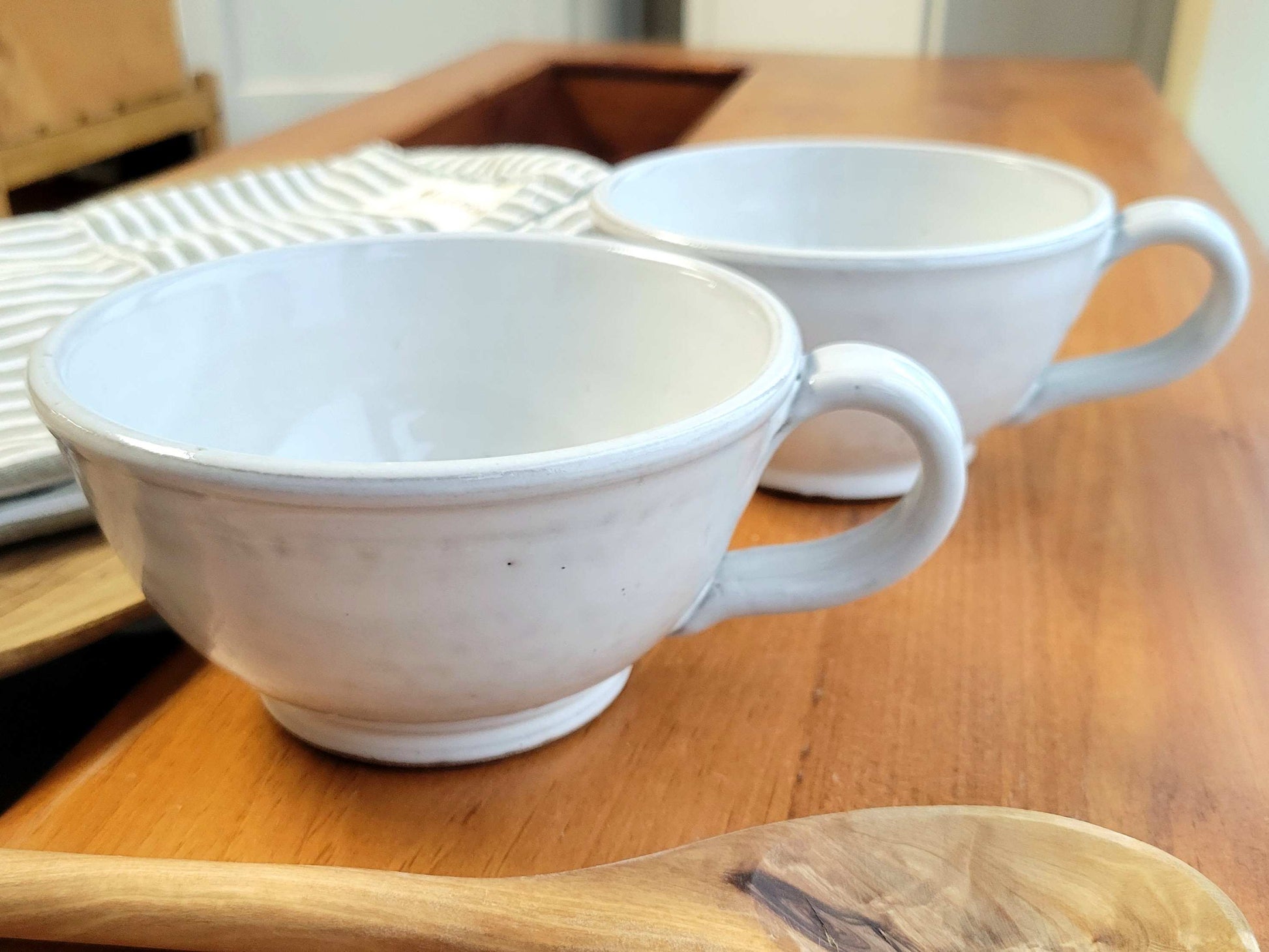 Two white ceramic handmade soup bowls with handles on a wooden table, with a striped napkin and a wooden spoon in the background.