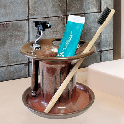 A brown or rust ceramic toothbrush stand with multiple slots, displaying a toothbrush, toothpaste tube, and a razor. The stand has a striped pattern and is placed on a light-colored surface. Great for bathroom organization. 