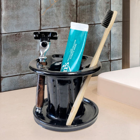 A black ceramic toothbrush stand with multiple slots, displaying a toothbrush, toothpaste tube, and a razor. The stand has a striped pattern and is placed on a light-colored bathroom counter. Stylish bathroom countertop organizer.