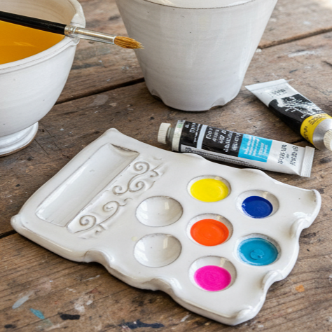 White ceramic palette with colorful paint wells on a wooden surface with paint tubes and a brush.