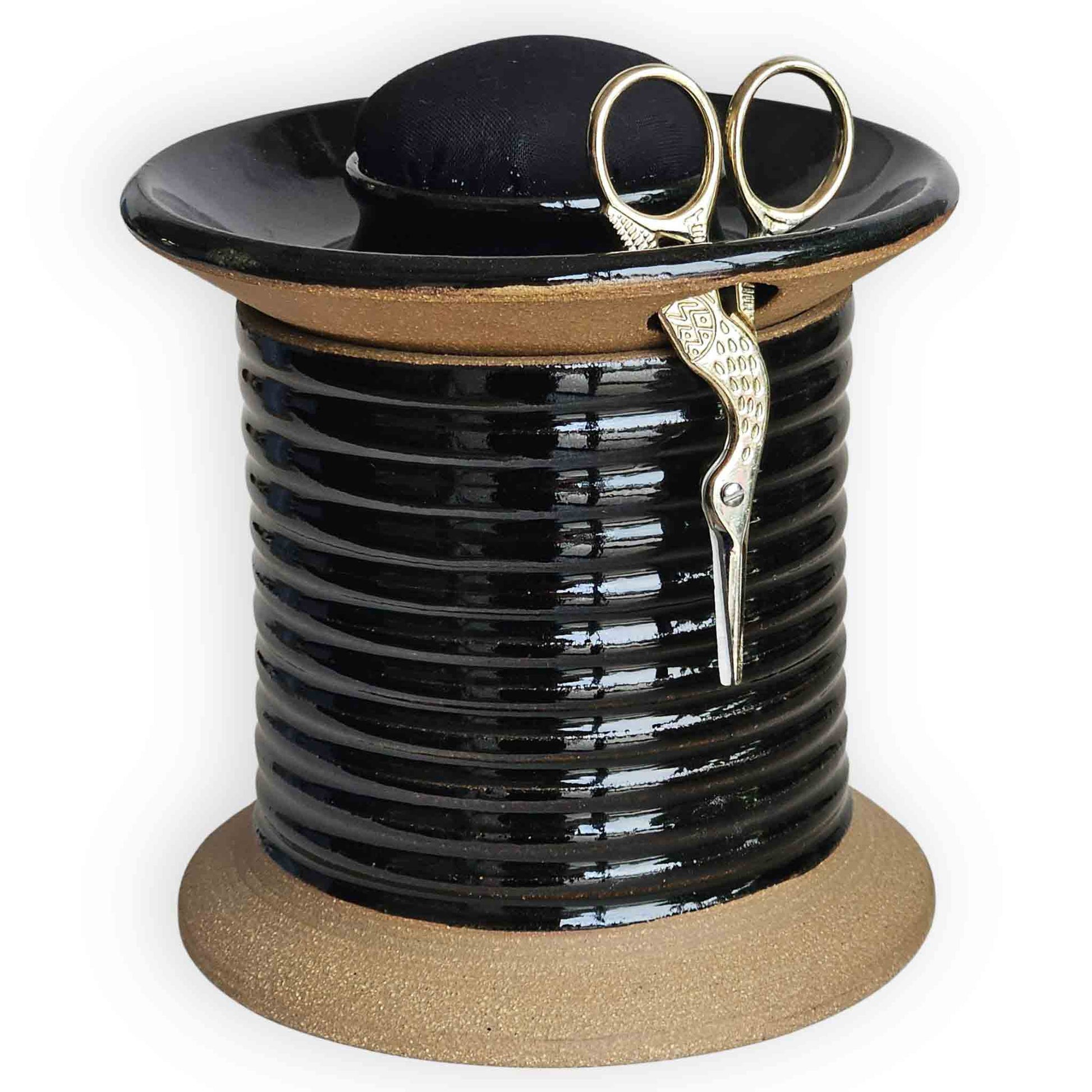 Mens sewing tool, an oversized spool-shaped black ceramic sewing station caddy with plain black pincushion topper, gold embroidery scissors and additional storage for tailors' tape and sewing notions.