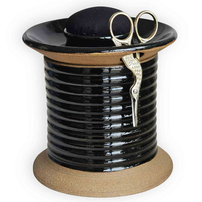 Mens sewing tool, an oversized spool-shaped black ceramic sewing station caddy with plain black pincushion topper, gold embroidery scissors and additional storage for tailors' tape and sewing notions.