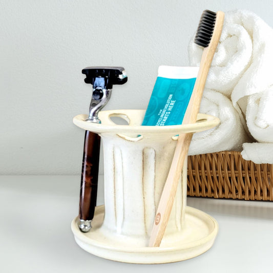 An off-white ceramic toothbrush stand with multiple slots, displaying a toothbrush, toothpaste tube, and a razor. The stand has a striped pattern and is placed on a light-colored surface with basket of towels beside it. Great for bathroom organization. 