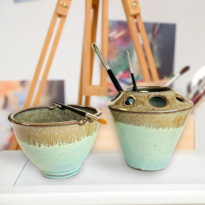 Handmade ceramic paint brush caddy and matching watercolor rinse bowl with multiple tool slots and brush rests, holding paintbrushes in Green and bronze tones. The pottery set is displayed in an artist’s studio with an easel and blurred artwork in the background.