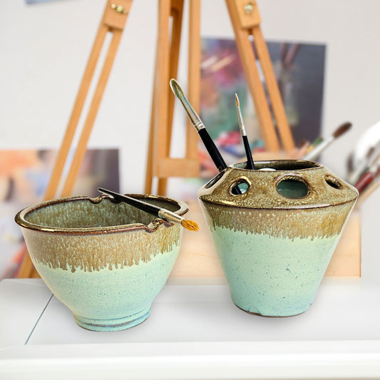 Handmade ceramic paint brush caddy and matching watercolor rinse bowl with multiple tool slots and brush rests, holding paintbrushes in Green and bronze tones. The pottery set is displayed in an artist’s studio with an easel and blurred artwork in the background.
