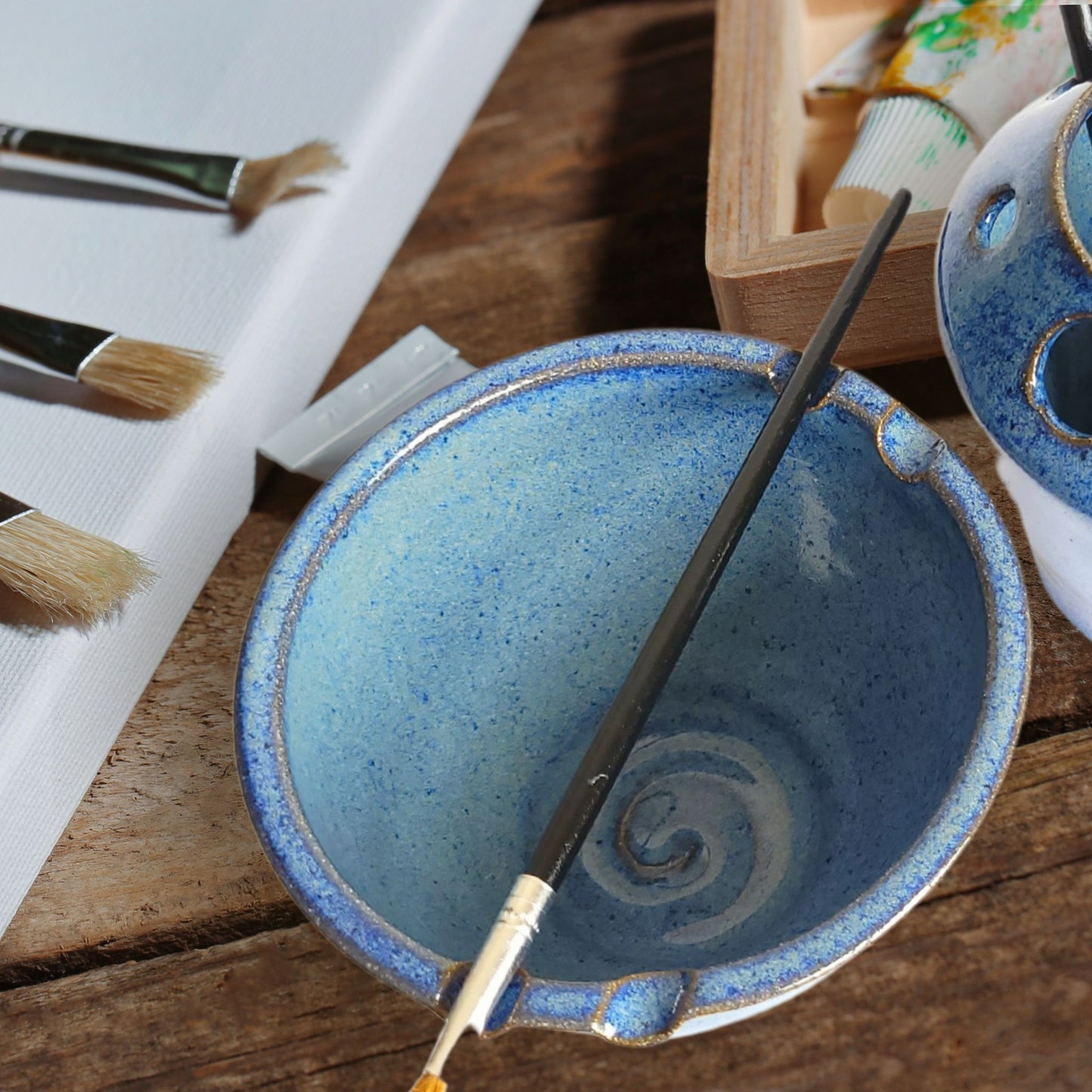 Unique handmade pottery brush rinse bowl with two side brush rests and functional swirl interior. Creative gift for artists, painters, and crafters set on wood table with painting project. 