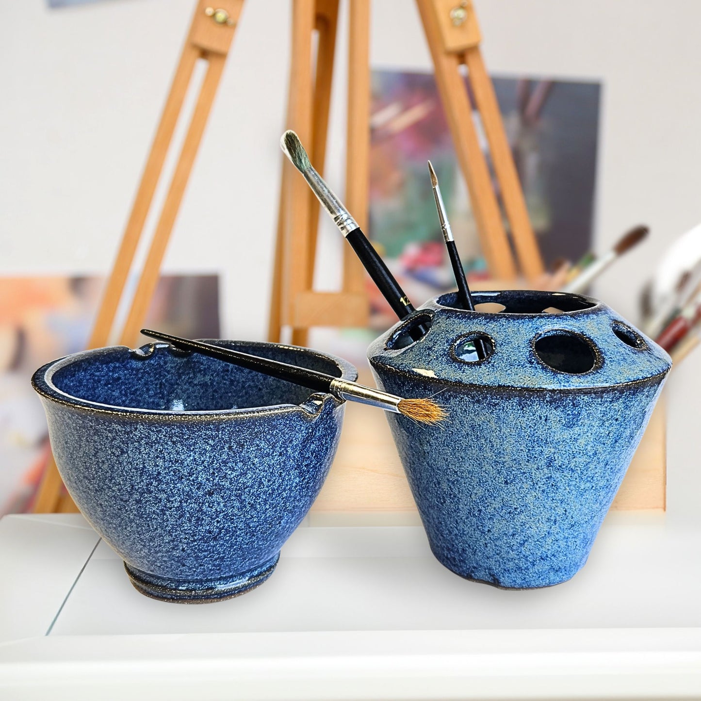 Handmade pottery artists brush holder and matching paint brush rinse bowl with multiple storage slots and brush rests, holding paintbrushes in speckled blue tones. The pottery set is displayed in an artist’s studio with an easel and canvases and blurred artwork in the background.