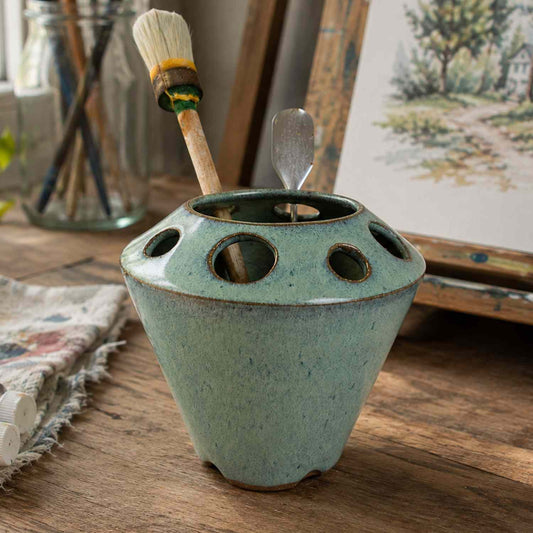 A paintbrush holder with circular cutouts, in a light green glaze, sitting on a wooden table. A paintbrush and a metal palette knife are placed inside the holder, with a rustic art studio background.