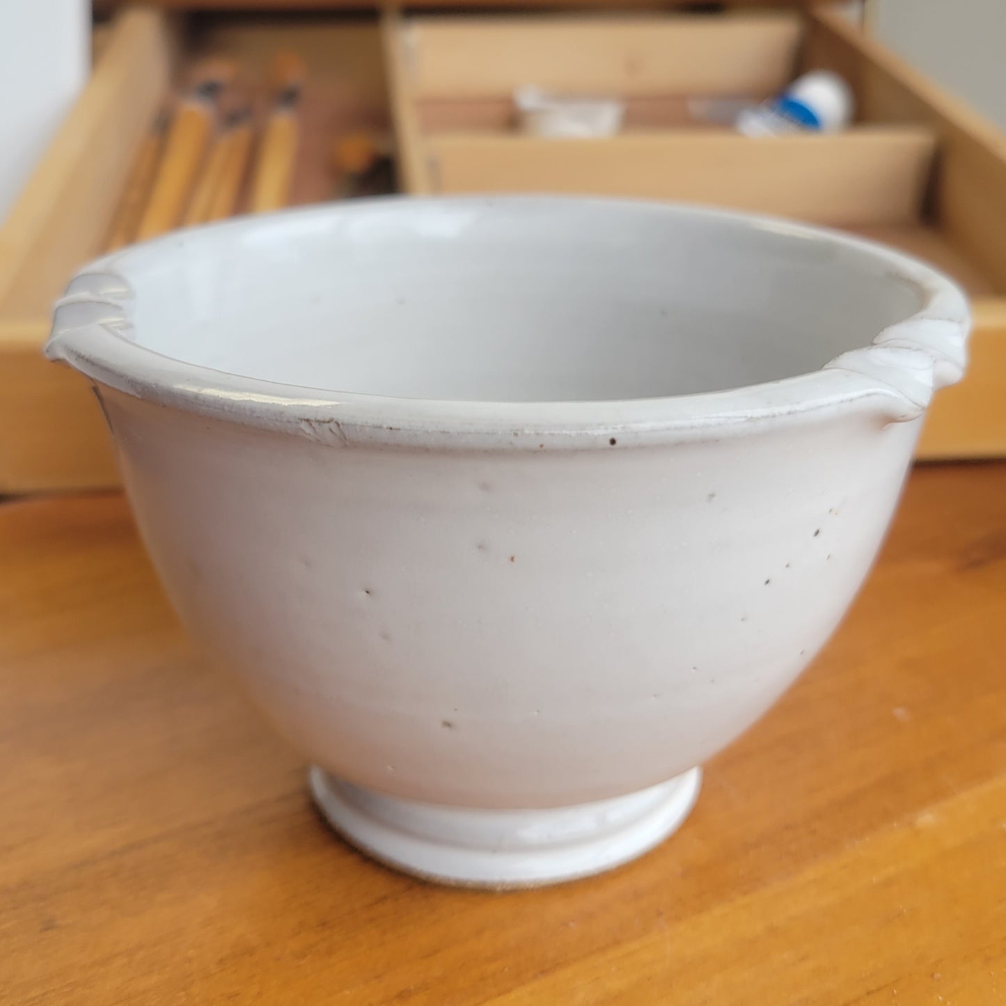 White ceramic painters brush rinse bowl on a wooden surface with blurred background and paint brushes and tubes of paint in background