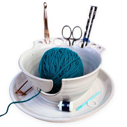 A ceramic yarn storage bowl with slots for tools such as scissors and needles, containing a ball of blue yarn and various crafting tools like a measuring tape and hooks.