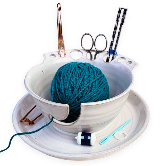 A ceramic yarn storage bowl with slots for tools such as scissors and needles, containing a ball of blue yarn and various crafting tools like a measuring tape and hooks.