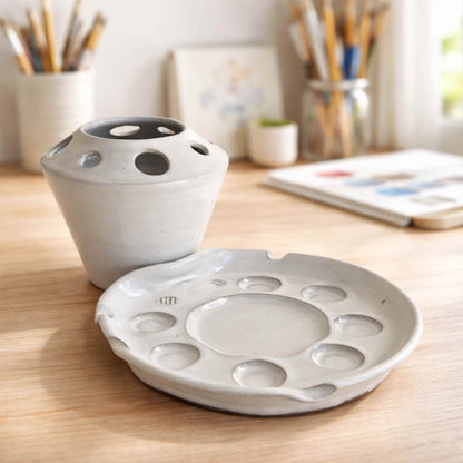 Handmade white ceramic paint palette with matching brush holder, featuring multiple brush slots and paint wells, styled on a light wood table in a bright artist studio