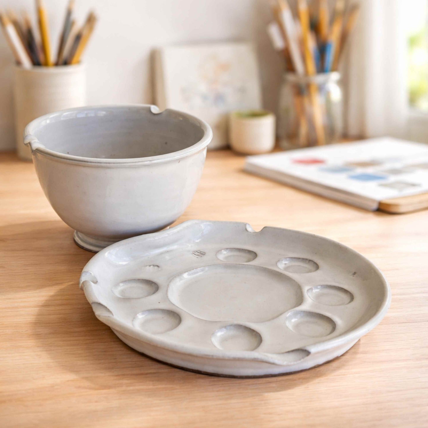 Handmade ceramic paint palette with rinse bowl for watercolor painting, featuring multiple paint wells and a smooth mixing surface on a light wood table in a bright studio