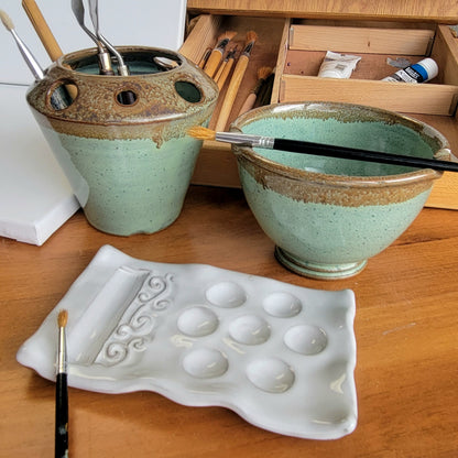 A handcrafted ceramic painting set including a palette, rinse cup, and brush caddy, displayed on a wooden surface with a potter's wheel in the background.