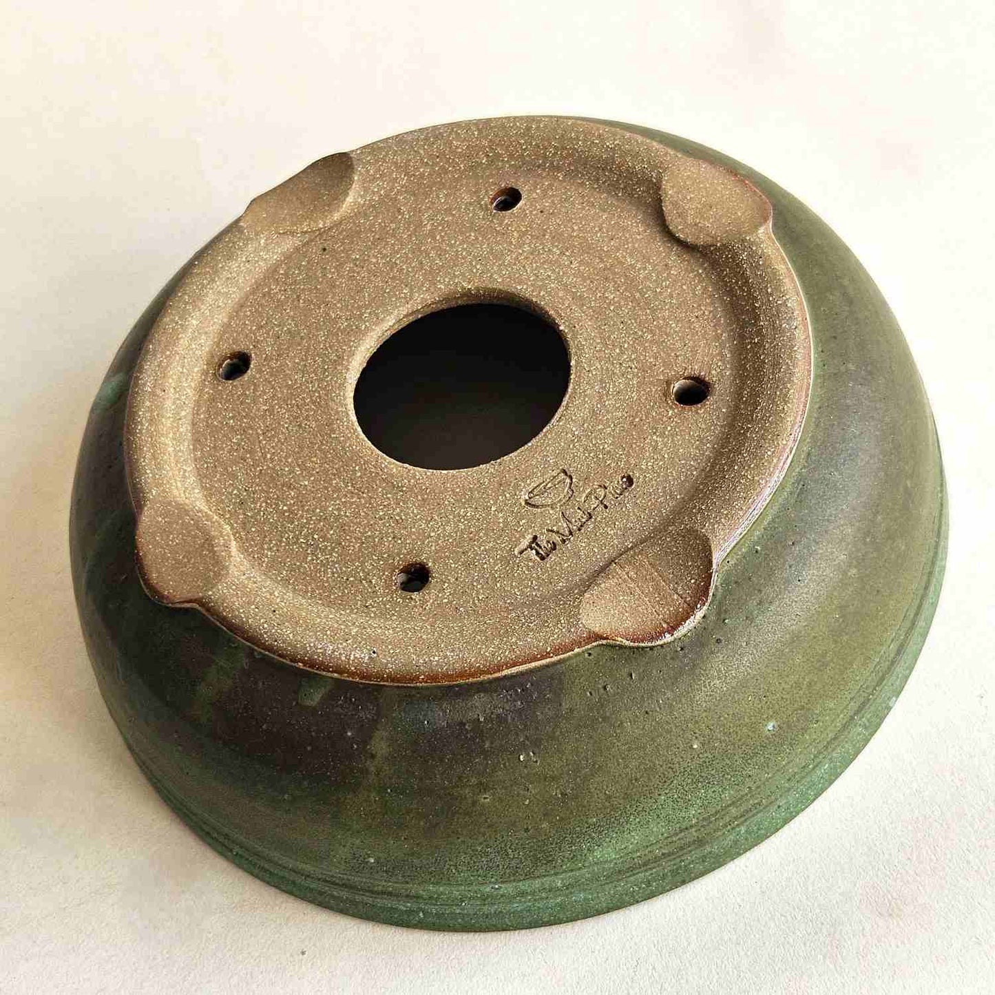 Bottom of a round shohin bonsai pot showing one drainage hole, four tie-down holes, and indented feet.