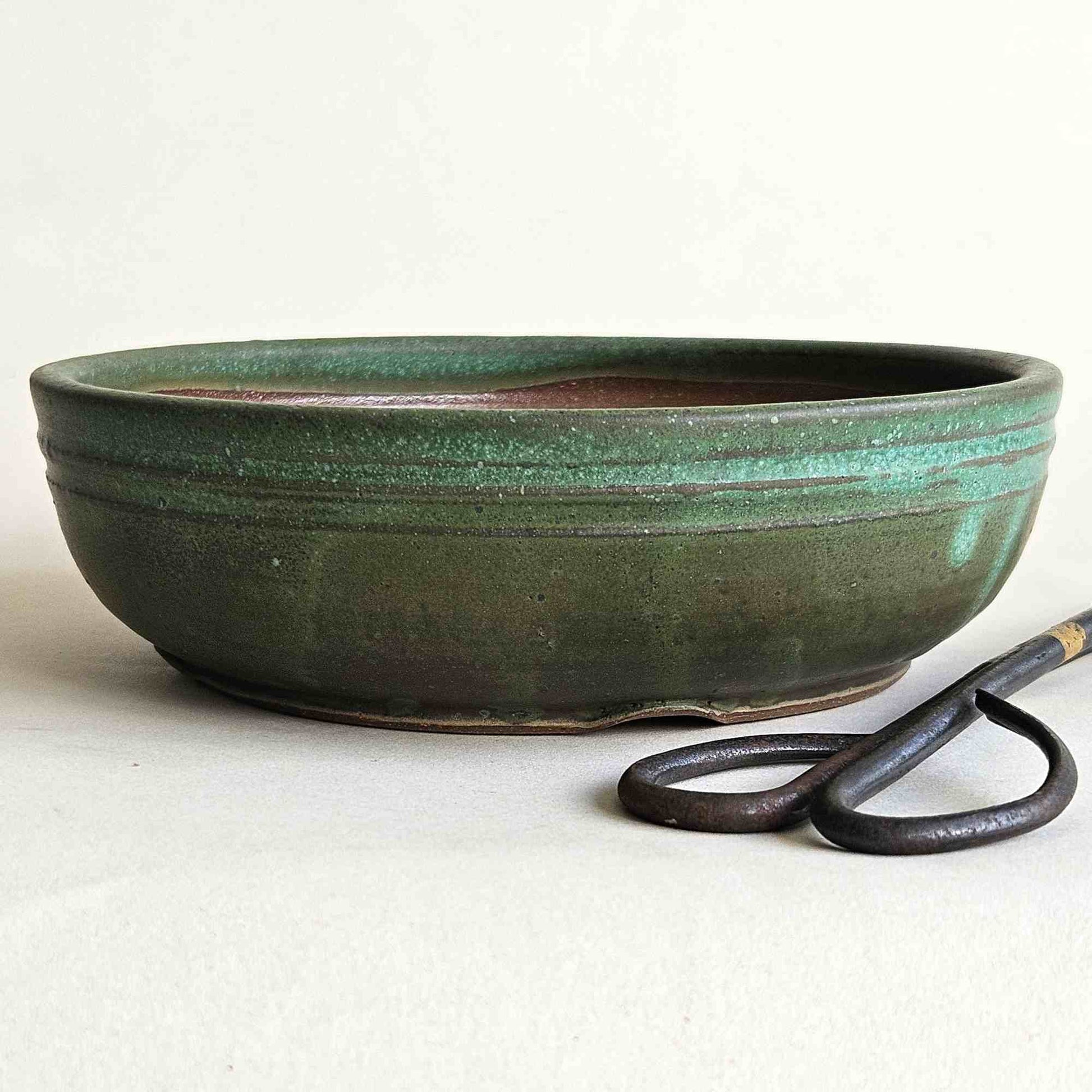 Low round green bonsai pot with a straight rim and subtle indented foot design, sitting next to bonsai scissors.