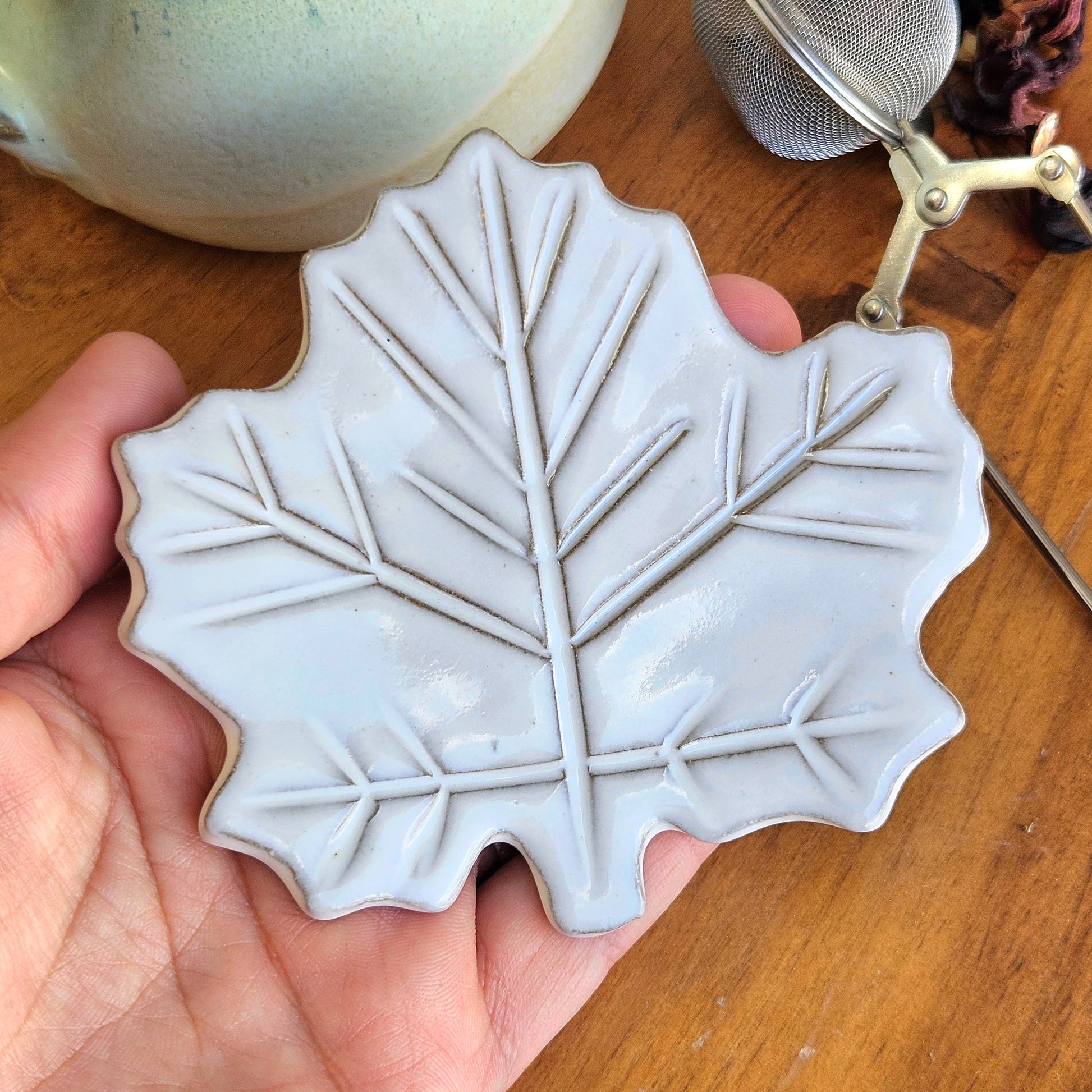 Handmade white maple leaf pottery teaspoon rest held in hand showing glaze texture with tea ball and loose tea with mug in background