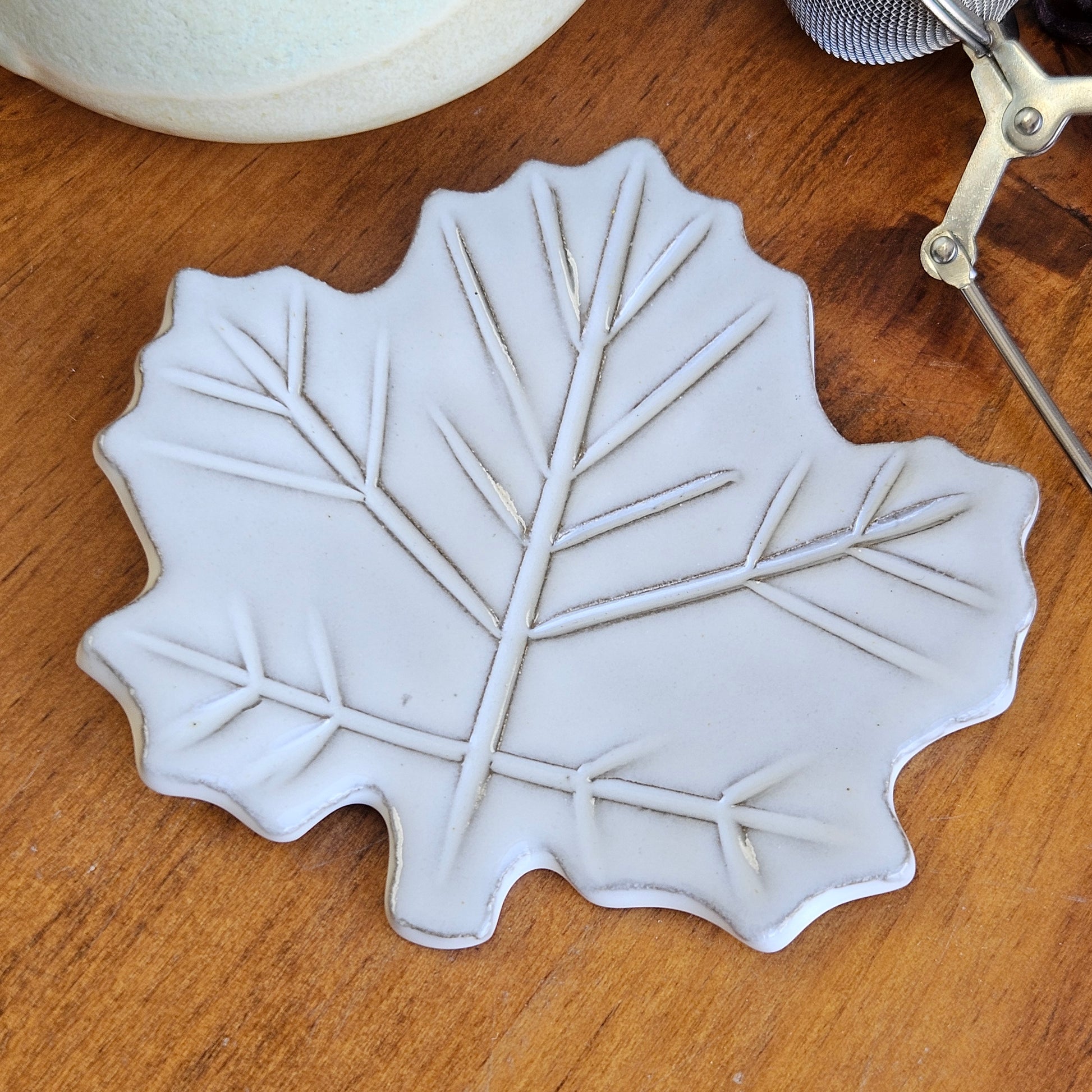 Mini maple leaf teaspoon rest in white ceramic on wooden surface beside teacup and tea ball