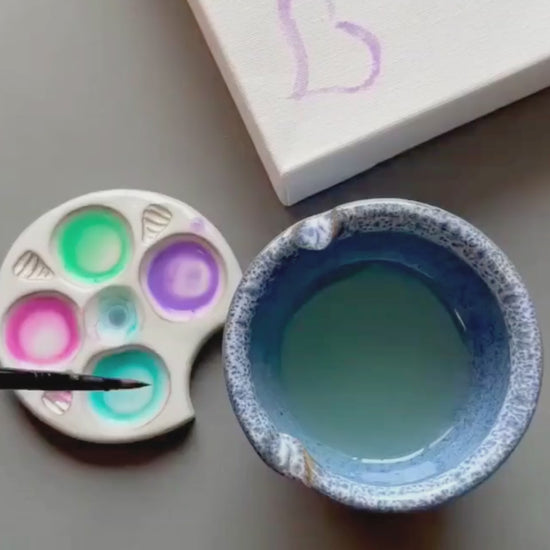 Top View of paint palette and rinse bowl with person painting hearts on a canvas in soft pastel watercolors
