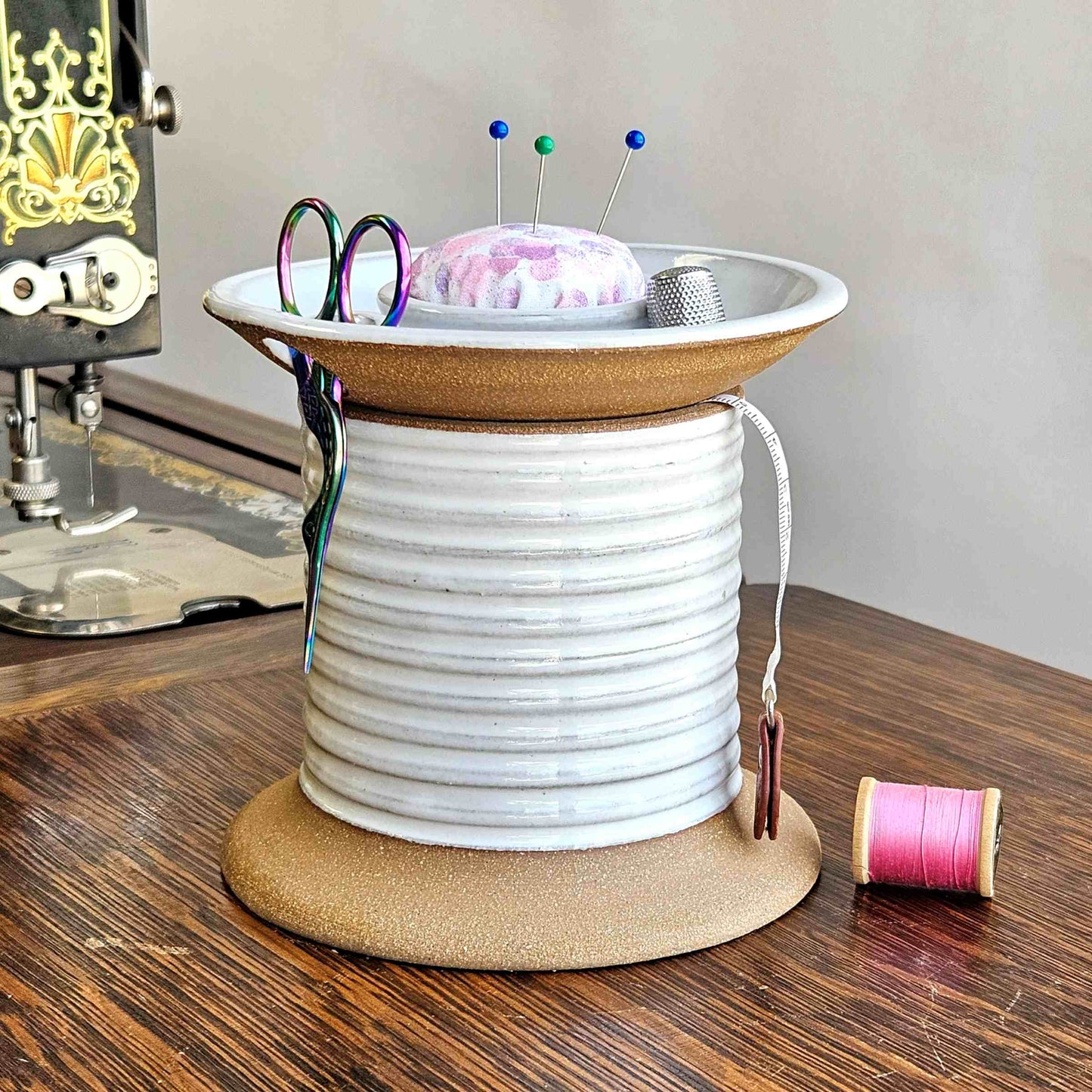 A ceramic sewing caddy with a farmhouse white color, resembling an oversized spool of thread. It has a removable lid with a slot for holding scissors and bobbin storage, and a fabric-covered pincushion on top.