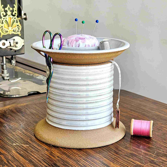 A ceramic sewing caddy with a farmhouse white color, resembling an oversized spool of thread. It has a removable lid with a slot for holding scissors and bobbin storage, and a fabric-covered pincushion on top.