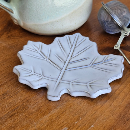 Small white maple leaf ceramic dish beside tea infuser and mug on wood surface