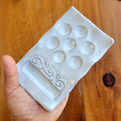 hand holding ceramic paint palette in white with 8 wells and mixing area and decorative swirl design on wooden background