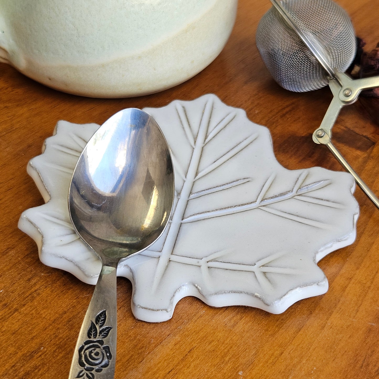 White mini maple leaf ceramic teaspoon rest holding silver spoon on wood table next to a ceramic mug and tea ball.
