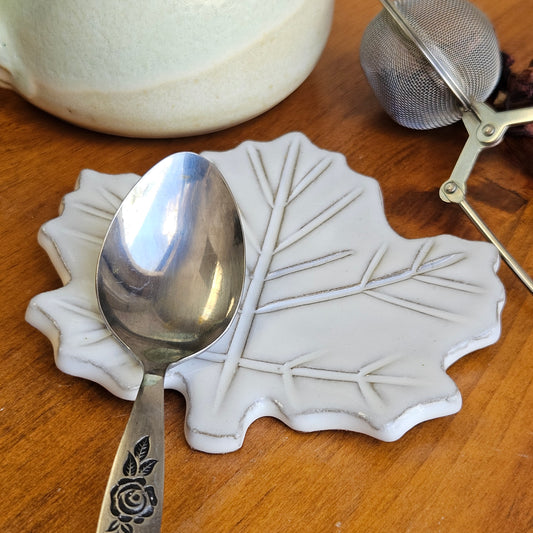White mini maple leaf ceramic teaspoon rest holding silver spoon on wood table next to a ceramic mug and tea ball.