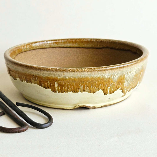 A shallow round ceramic bonsai-style pot with a brown and beige dripping glaze, shown on a plain light background with a pair of pruning shears partially visible in the lower left.