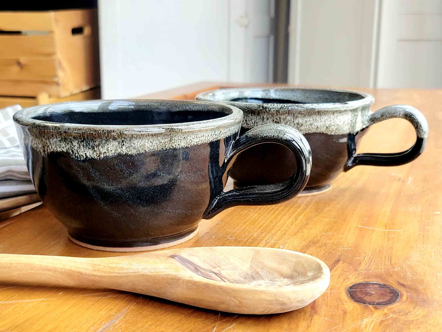 Two black ceramic soup bowls with white speckles and handles, placed on a wooden surface with a wooden spoon beside them.