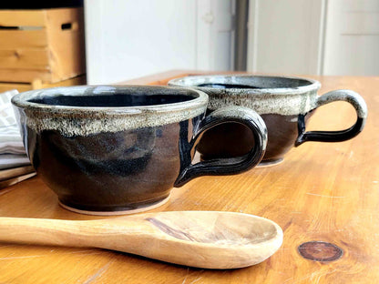 Two black ceramic soup bowls with white speckles and handles, placed on a wooden surface with a wooden spoon beside them.