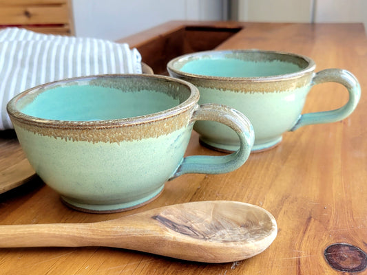 Two ceramic soup bowls with handles in green and bronze colors, displayed on a wooden table with a wooden spoon beside them, perfect for soups, stews, and pasta.