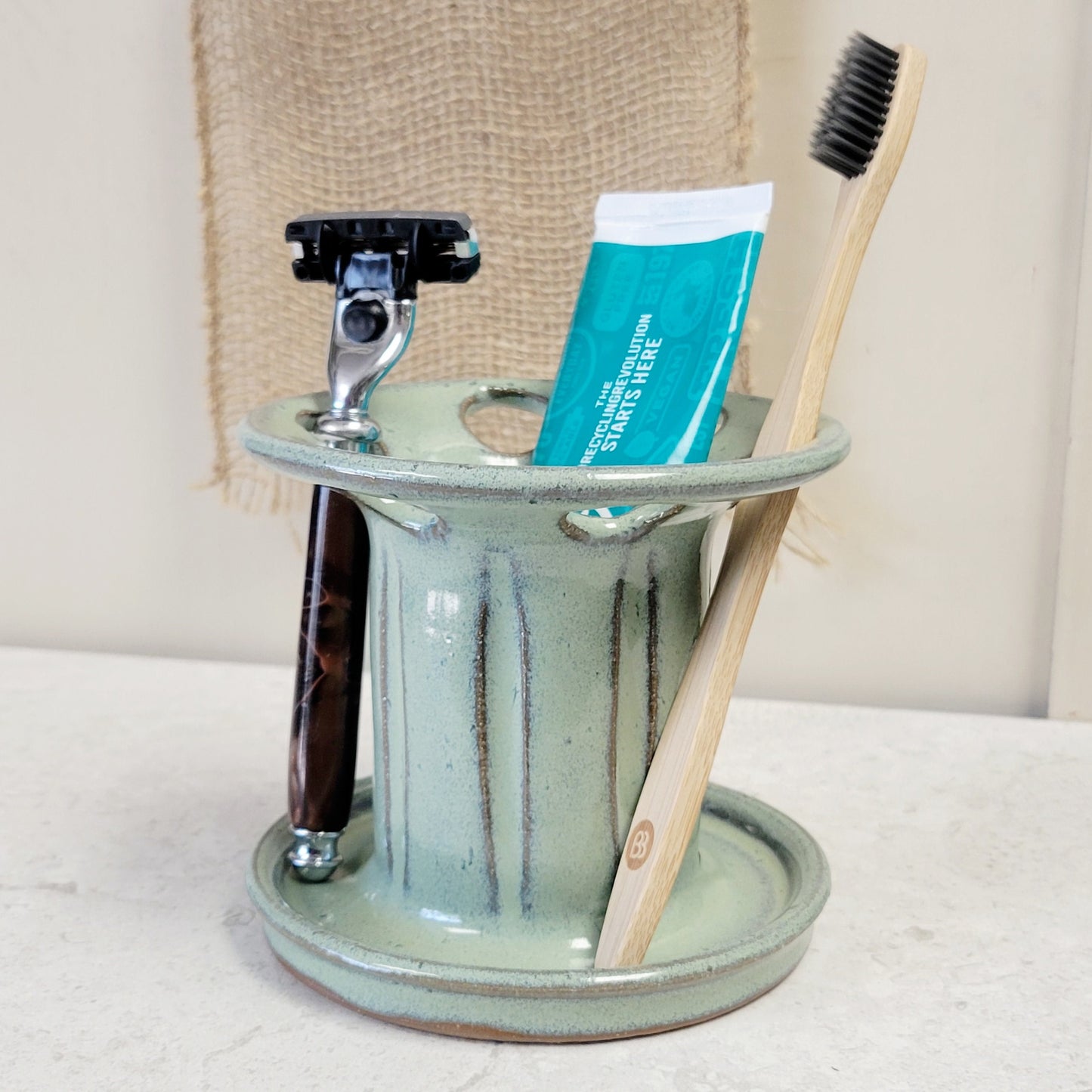 A handmade green pottery toothbrush holder with multiple slots, containing a toothbrush, toothpaste tube, and a razor.
