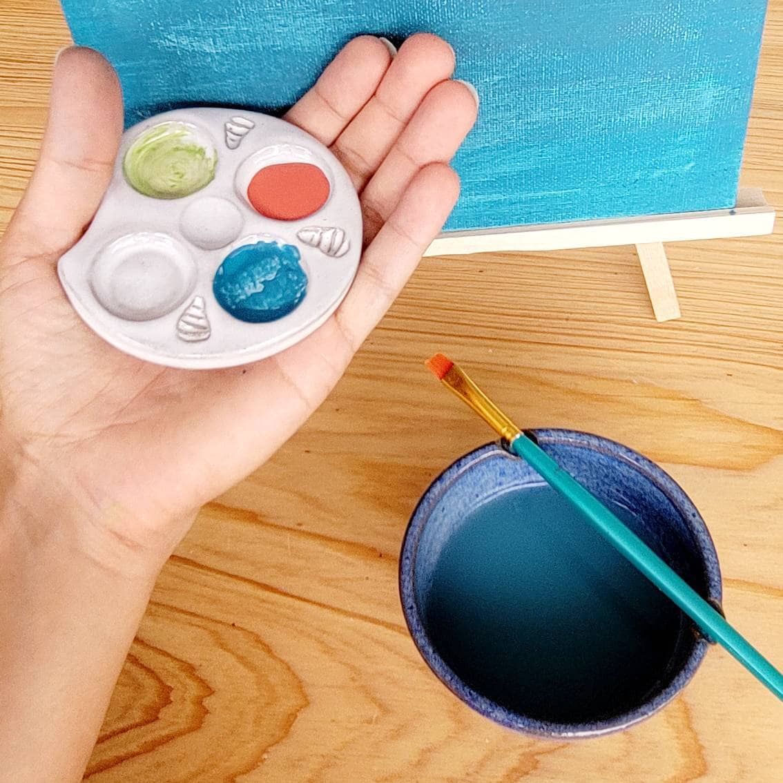 Hand holding a small white ceramic watercolor palette filled with paint beside a blue ceramic rinse bowl on a wooden table.