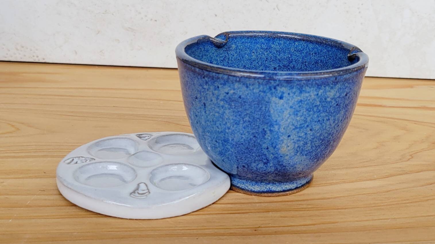 Blue ceramic rinse bowl with built-in brush rest and matching white ceramic paint palette displayed on a wooden surface.
